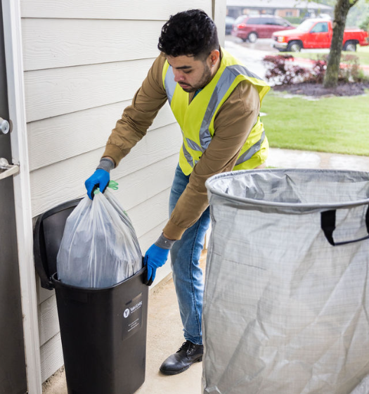 A person wearing a yellow safety vest and blue gloves places a tied trash bag into a black outdoor garbage bin beside a house, with a large gray trash container nearby and a lawn with a red vehicle in the background.
