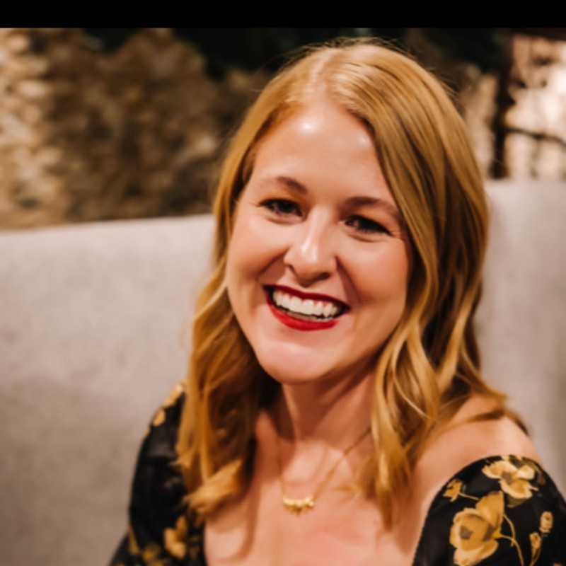 A woman with shoulder-length blonde hair smiles warmly. She is wearing a black dress with a floral pattern and a gold necklace, seated indoors with a softly blurred background.