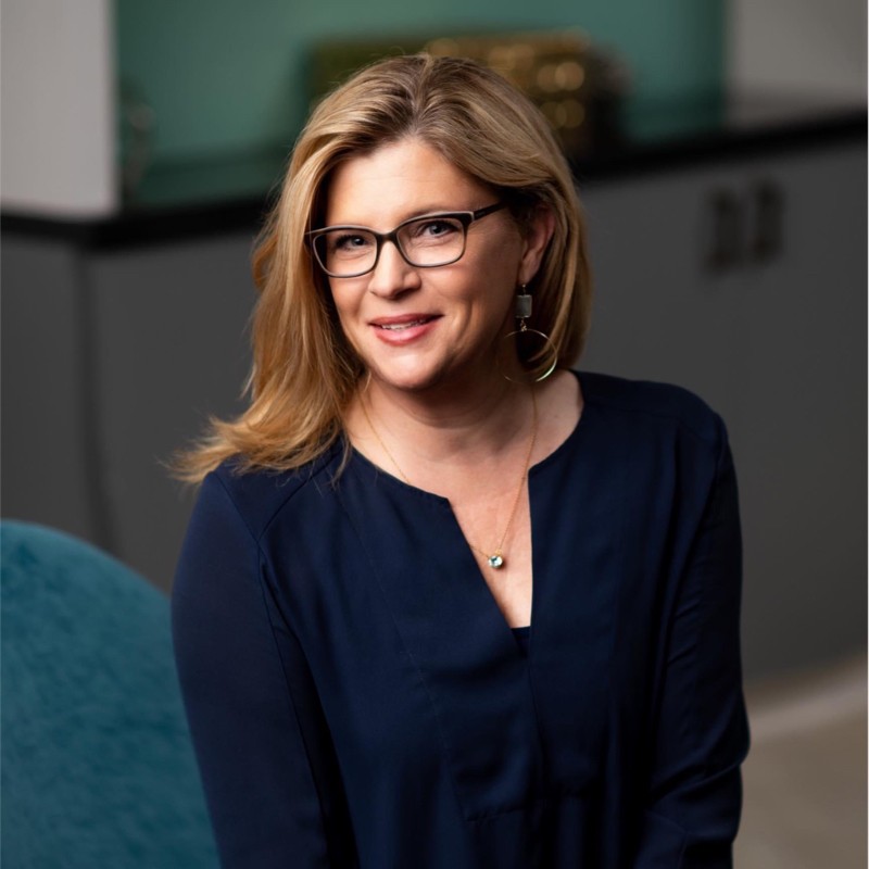 A woman with blonde hair and glasses smiles at the camera. She is wearing a dark blue blouse, a necklace, and large hoop earrings, sitting indoors with a blurred background featuring cabinets.