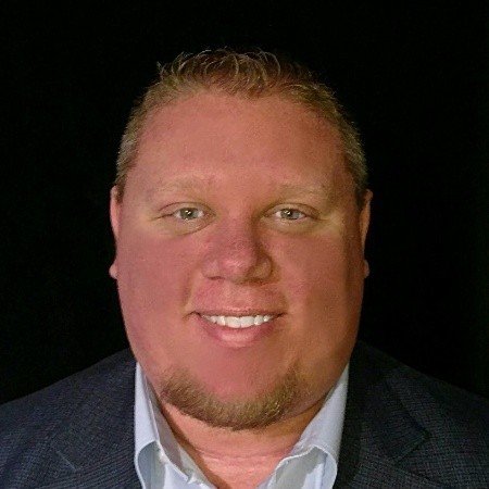 A man with short light brown hair, light eyes, and a trimmed goatee smiles at the camera. He is wearing a dark blazer over a light-colored collared shirt, with a dark background behind him.