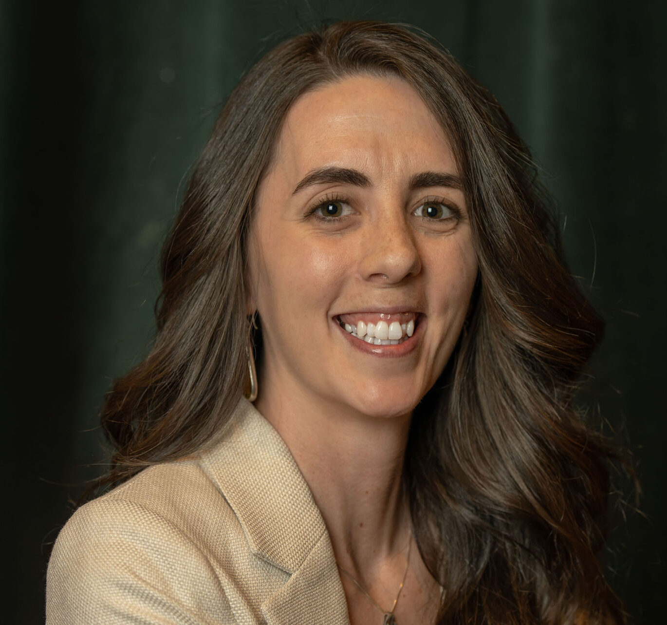 A woman with long brown hair and a beige blazer smiles at the camera, standing in front of a dark green backdrop.