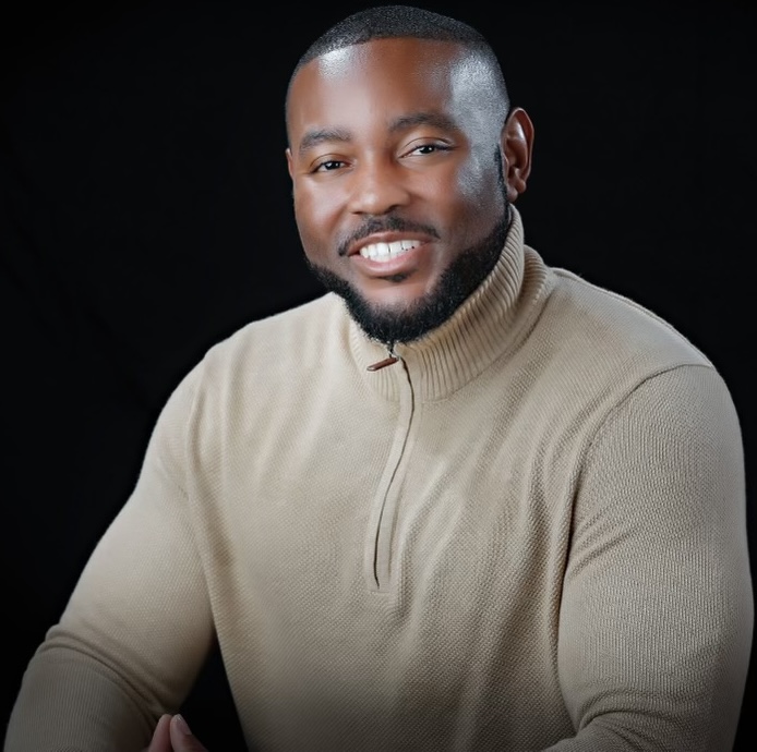 A man with a short beard and close-cropped hair smiles at the camera. He is wearing a beige, long-sleeve, zip-up sweater and is posed against a solid black background.