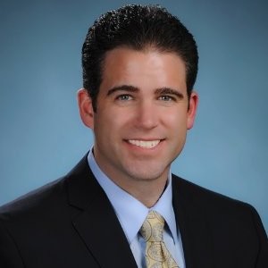 Professional headshot of a man with short dark hair, wearing a dark suit, light blue shirt, and patterned yellow tie, smiling in front of a plain blue background.