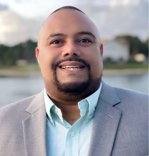 A smiling man with a trimmed beard, wearing a light gray blazer and a light blue shirt, stands outdoors with a blurred background of water and trees.
