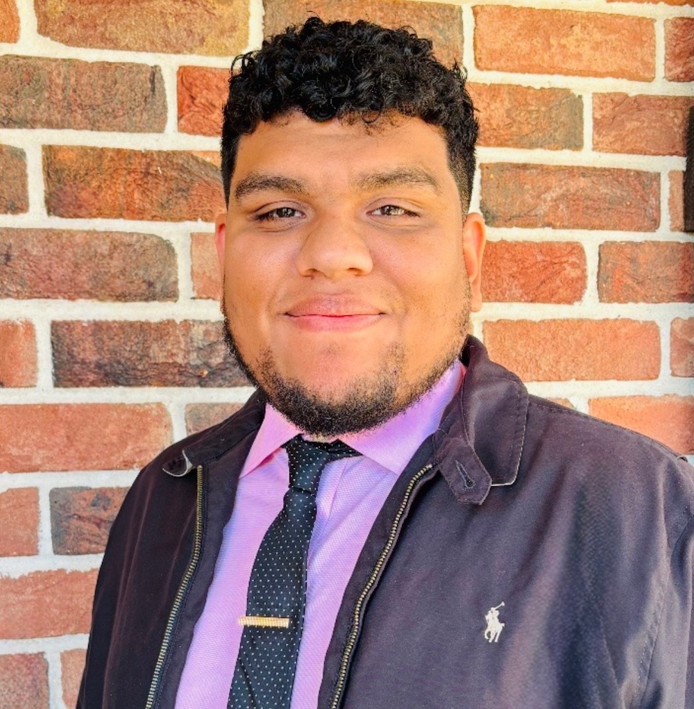 A man with short curly hair and a trimmed beard smiles at the camera. He is wearing a black jacket, a purple shirt, and a black tie with small white dots. Behind him is a brick wall.