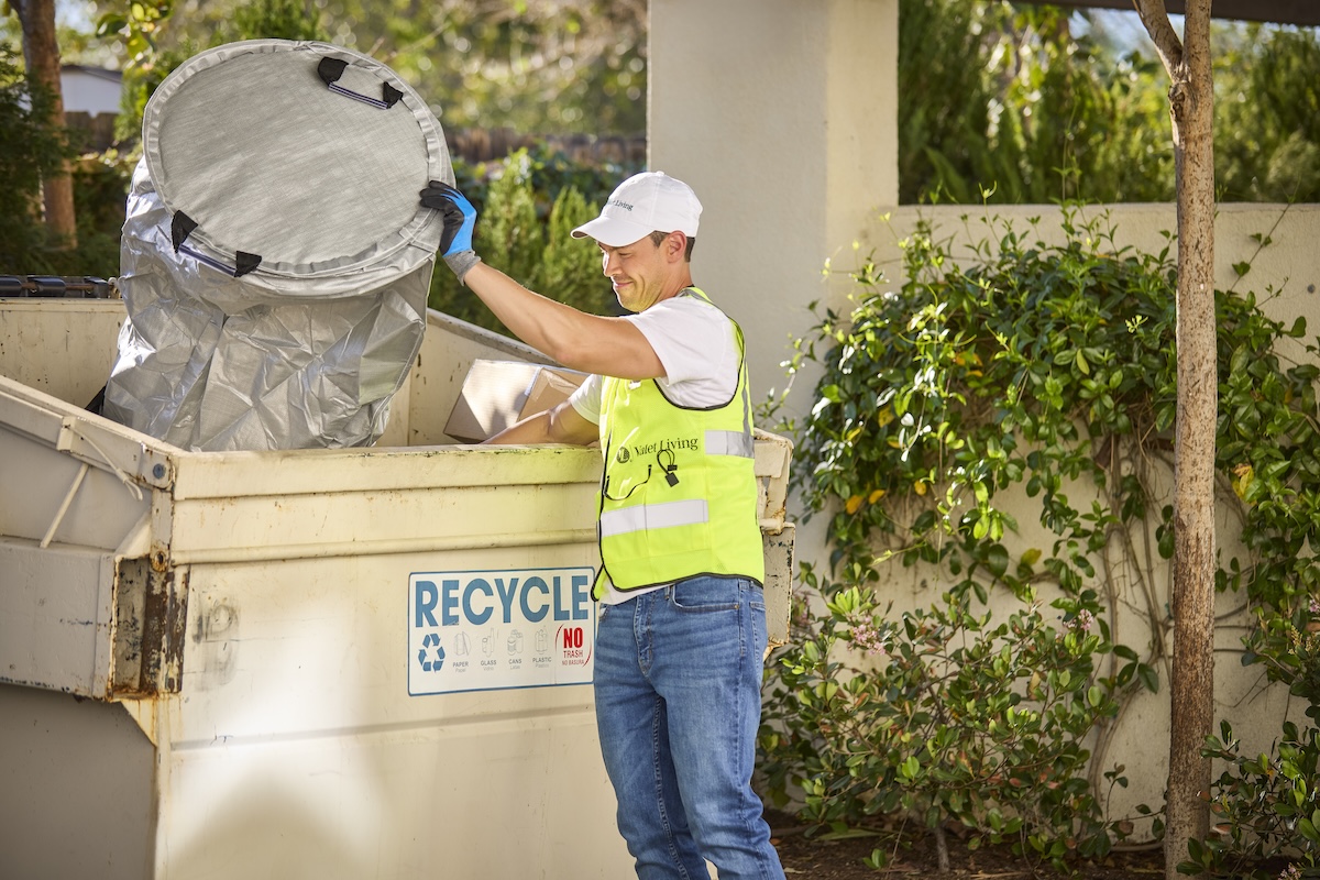 A person wearing a white cap and safety vest places a large trash can into a recycling dumpster outdoors, surrounded by greenery.