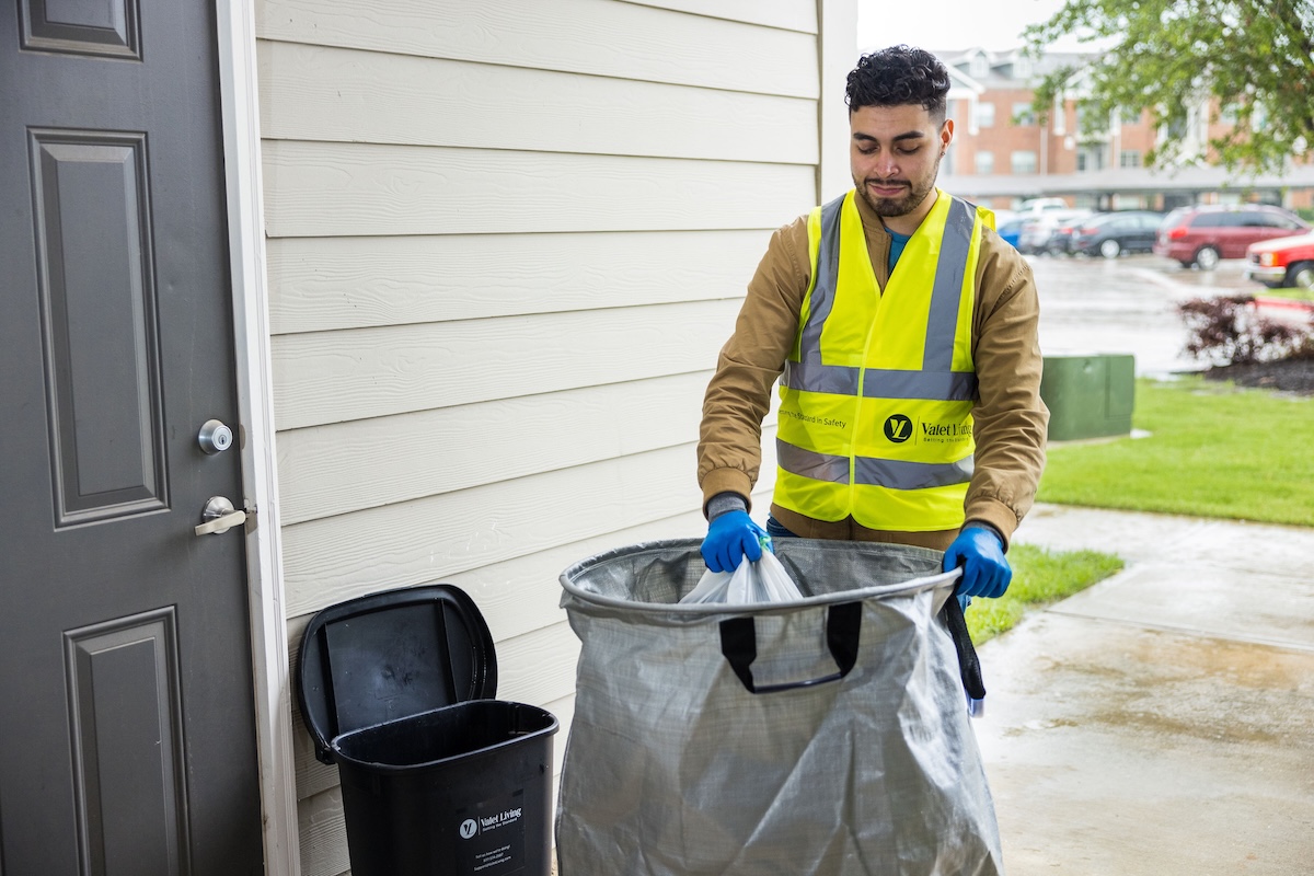 A man in a yellow safety vest and gloves places trash into a large bag outside near a black bin on a rainy day.